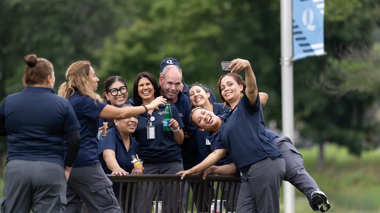 A group of facilities workers in Quinnipiac uniforms smile excitedly for a selfie photo outside