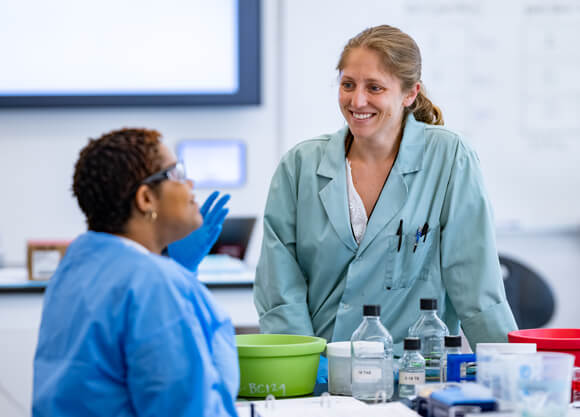 A faculty member in a blue lab coat speaks with a student with lab equipment on the table